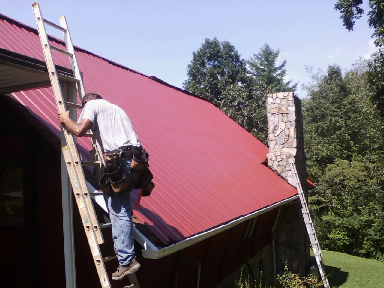 Roofer on a ladder beside a red metal roof and stone chimney — Soffit & Fascia Repair by Roofing Brothers in Tustin